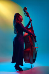 Young African-American talented woman, musician playing on double bass in neon light against...