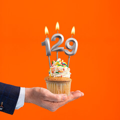 The hand that delivers cupcake with the number 129 candle - Birthday on orange background