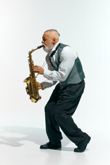 Naklejka premium Jazzy vibes. Bearded musician in sleeveless gray vest and black pants plays saxophone energetically against white studio background. Concept of music, festivals, contemporary art, hobby, creativity.