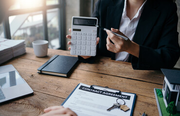 Business meeting showing a person using a calculator and signing a contract agreement at a wooden desk in an office environment.