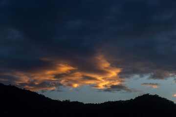 Landscape of thick clouds, orange light hitting the blue sky, silhouettes of trees, forests, and mountains. Area for text in the middle of the image