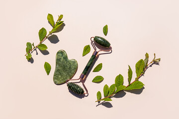 Green malachite roller massager and jade gua sha scraper on a beige background with green natural twigs. top view. flat lay. Natural care.