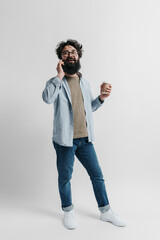 Fototapeta premium A cheerful bearded man in casual attire is talking on a smartphone and holding a takeaway coffee cup against a plain background. He conveys a vibe of casual business and approachability.
