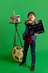 Exited, little child, boy dressed as film director posing holding clapperboard against green studio...