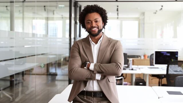 A confident professional in a taupe suit leans against a white table in a bright office setting, arms crossed and smiling at the camera. The concept captures a relaxed yet assured business atmosphere