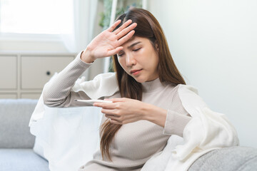 Sick, influenza asian young woman, girl headache have fever, hand touching forehead, holding thermometer for check measure body temperature, illness while sitting rest on sofa at home. Health care.