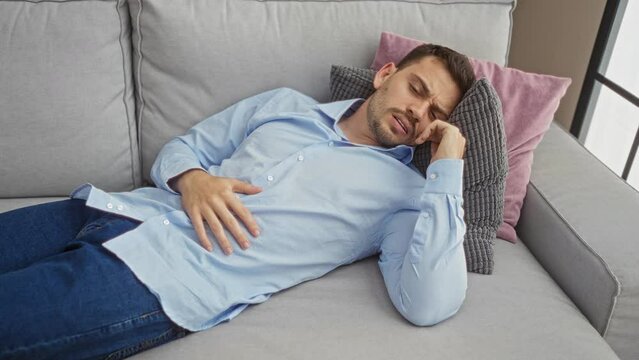 Young man with a beard in a blue shirt and jeans lies on a couch holding his stomach in a living room, appearing to be in pain.