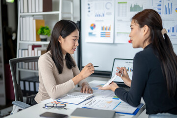 Two women are sitting at a desk with a whiteboard behind them