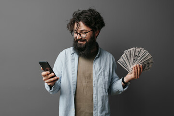 Smiling, bearded man in casual clothes looks at his smartphone screen while holding a fan of cash...