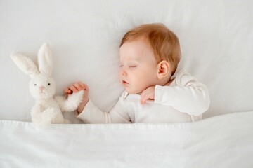 A baby boy sleeps under a blanket in a white bodysuit on the bed with his hand folded under his...