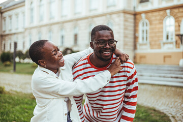 A smiling black male and female college students share a playful moment outside an academic building, dressed in casual attire suitable for a day of studies and relaxation.