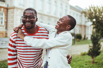 A delighted African American college couple laugh together outdoors, with the woman embracing the...