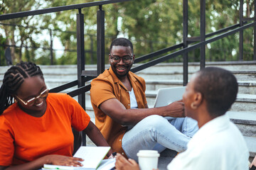 A group of black university students engages in a lively discussion on campus stairs, with books...