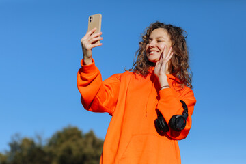 Happy young woman in orange sweatshirt holds headphones, uses smartphone for vlog, blog, waves hand...
