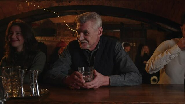 Three people drinks at the bar counter. Caucasian adult male enjoying a drink in restaurant. Addicted alcoholic order whiskey. Laughing man takes one more glass irish whisky.