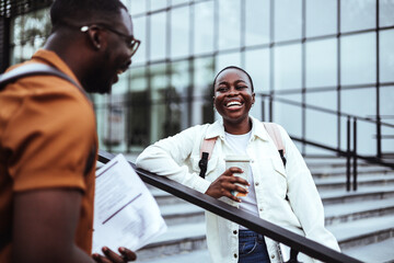 A joyful male and female student engage in a friendly conversation on university stairways, with books in hand, reflecting a casual educational setting.
