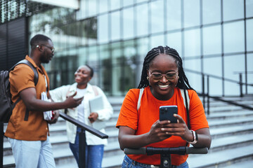 A radiant black female student in a vibrant orange shirt smiles while using her smartphone, standing near a university building with diverse classmates in the background.