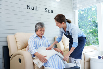 Fototapeta premium Asian girl, foot spa shop owner, preparing to serve and recommend services to customer. Girl in denim apron assists elderly customer with blanket, showcasing care, professionalism, and attention.