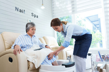 Obraz premium Asian senior woman receiving pedicure at nail spa from young Asian female staff. Cozy spa setting with bright natural light. Woman appears relaxed, enjoying professional foot care service.