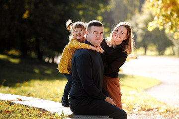 Fototapeta premium Family day. Happy young mom, dad and little child daughter having fun, kid bits fathers ear at autumn park, enjoying spend time together at weekend. Concept of parental care, happy carefree childhood