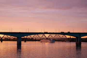 A four-deck cruise passenger ship sails along the river under the bridge during sunset. Cruises and river trips