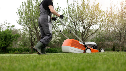 Professional gardener in protective apparel is mowing green grass lawn using modern gasoline...
