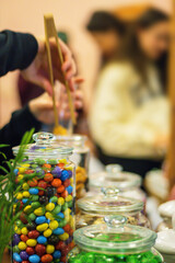 Young woman selling candies at the food market. She is picking out candy with wooden or bamboo tongs out of the glass jar and putting them into plastic bag. Copy space. Selective focus, Blurred hands