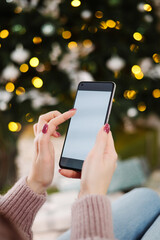 christmas mockup with space for text. christmas advertising, app template. hands holding phone with empty screen on background of beautiful christmas tree with lights