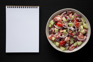 Homemade Chopped Mediterranean Salad in a Bowl, blank notepad on a black background, top view.