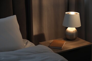 Stylish lamp and book on bedside table near bed indoors