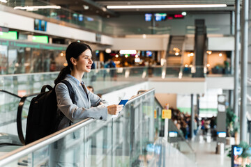 Woman holding passport, boarding pass and smartphone standing near window at airport lobby and waiting for departure. Tourist in airport is standing near panoramic window waiting for security check.