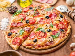 Pizza topped with cherry tomatoes, salami and other ingredients is on wooden table surrounded by ingredients. Top view.