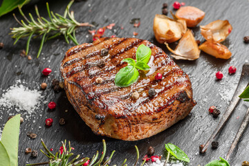 Grilled delicious ribeye steak and some seasonings and herbs on black slate serving plate. Flat lay.