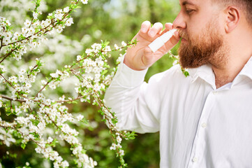 Man allergic using medical nasal drops, suffering from seasonal allergy at spring in blossoming garden. Handsome man treating runny nose in front of blooming tree outdoors. Spring allergy concept.