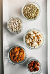 Bowls, nuts and seeds on plate, above and hazelnut for nutrition, health and table with diet for wellness. Cashew, almond and pumpkin in glass container for meal prep with food in morning at house