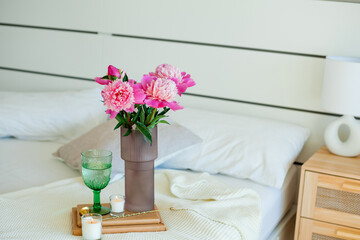 Bouquet of peonies in glass pink vase, green glass of water, candles on the bed. Bedroom. Spring flowers.
