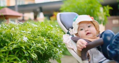 A happy baby wearing a sun hat sits in a stroller outdoors, surrounded by lush greenery and white...