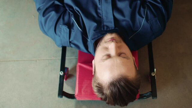 View from above of Caucasian bearder mechanic lying on creeper while repairing broken car. Busy man wearing special uniform while fixing detail with wrench. Auto maintenance concept.