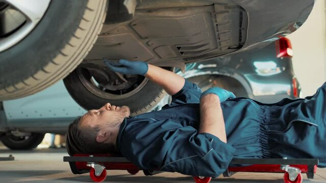 Side view of Caucasian engineer lying under car on roller. Busy man checking serviceability of parts. Worker repairing issue in automobile. Making diagnostics. Working in auto service.