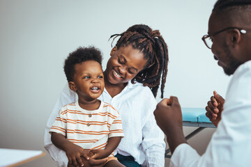 A joyful black boy sits on his mother's lap during a medical consultation as the doctor engages...