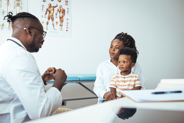 A young black boy sits attentively with his mother during a consultation as a doctor in a white...