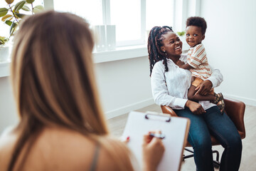 A joyful black boy sits on his mother's lap during a consultation with a healthcare professional in...