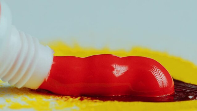 Bright glossy paint of red color being squeezed out of tube onto a yellow spot on paper. Extreme close-up shot