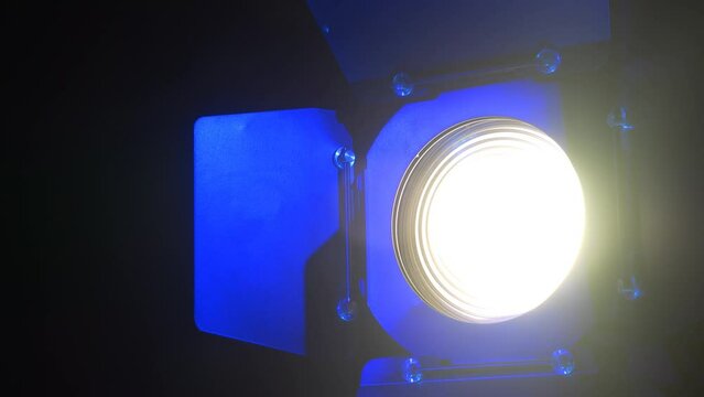 Theater spotlight with a fresnel lens with curtains on a black background with blue reflections, turning towards the camera. Medium plan