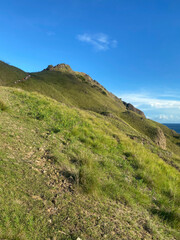 Stunning view of a grassy hill under a bright blue sky, with hikers in the distance ascending the trail. Ideal for nature, hiking, and outdoor adventure themes