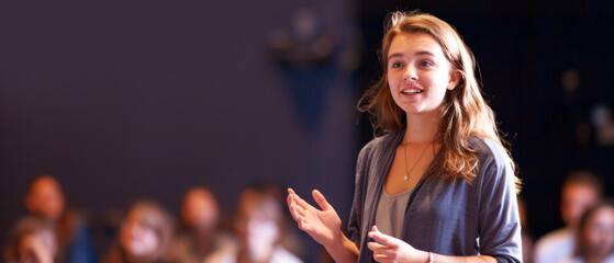 A large crowd is seen from behind as they attentively listen, with warm light illuminating the speaker at the front.