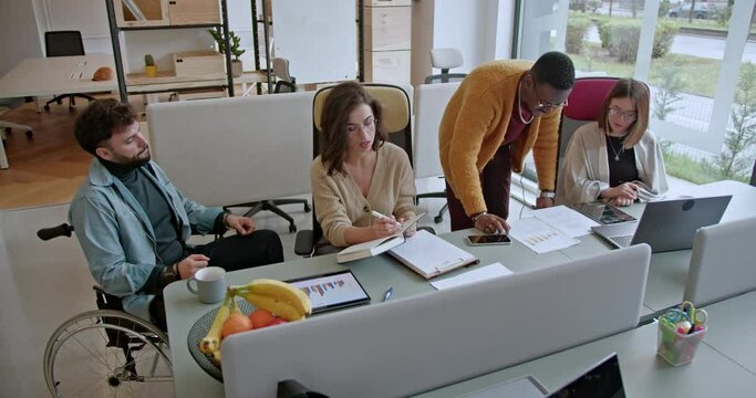A group of business employees, representing different ethnicities, is seen discussing and collaborating on a new project. They are surrounded by charts and laptops in a bright and modern office space.