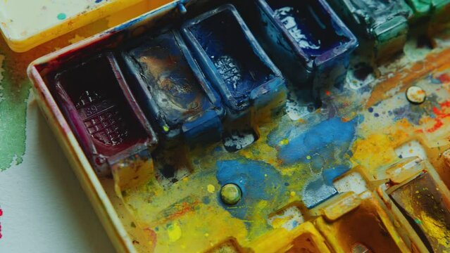 Watercolor paint set and couple of paintbrushes on paper with colorful stains. Extreme close-up shot