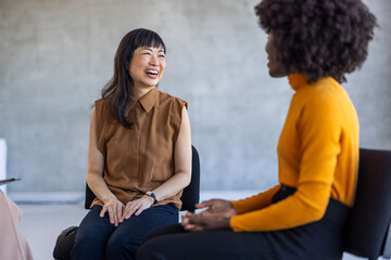 Two professional women, one Asian and one African ethnicity, laughing and enjoying a genuine connection at a modern office workspace.