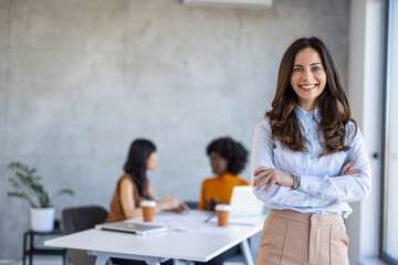 Professional female with a radiant smile stands in a contemporary workspace with colleagues interacting in the background.